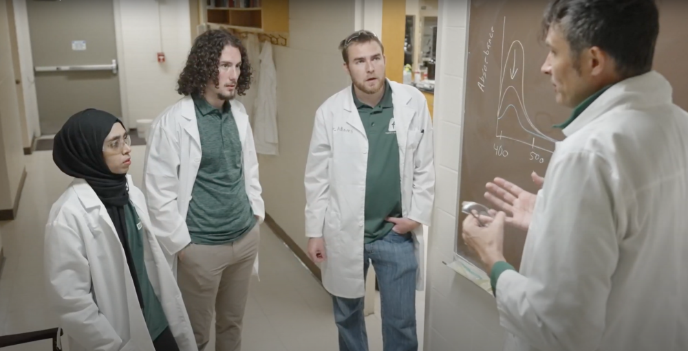 4 people wearing lab coats and Charlotte 49er gear talk in front of a chalkboard.